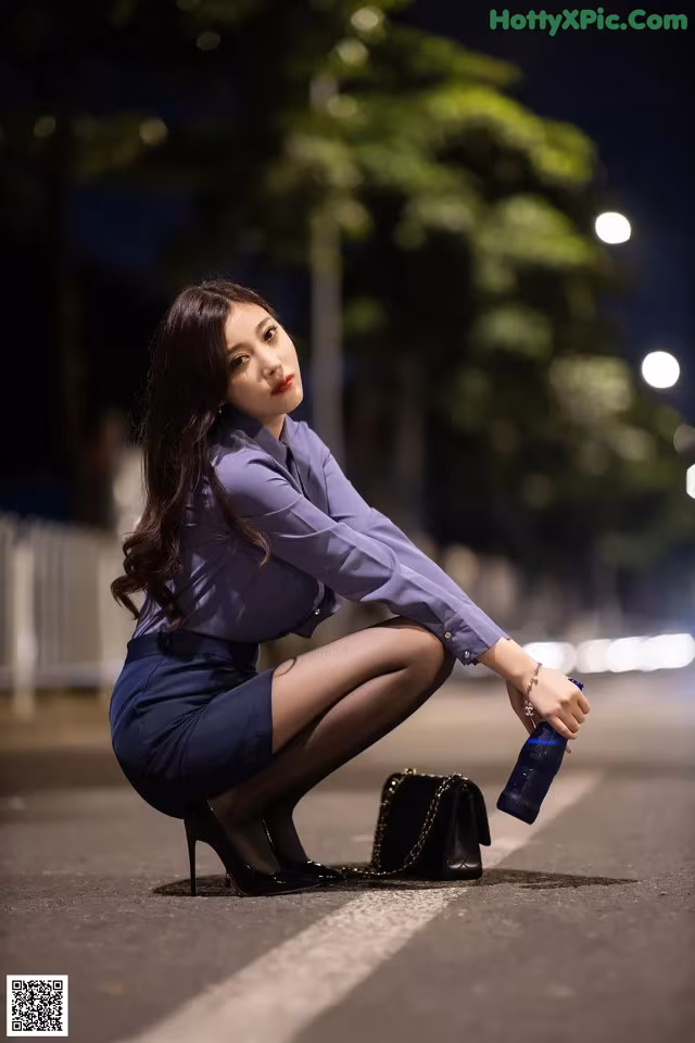 A woman in a blue shirt and skirt crouching down on the street.